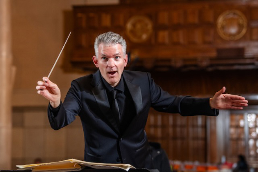 Frontal shot of Artistic Director Michael Driscoll in black tuxedo conducting with his baton.
