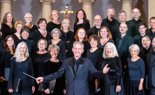 Artistic Director Michael Driscoll with arms outstretched in front of members of Cmomonwealth Chorale in all-black concert dress.